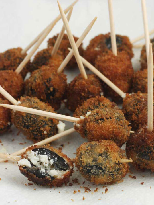 A plate of Greek fried olives with a cocktail stick in them on baking paper on a white plate