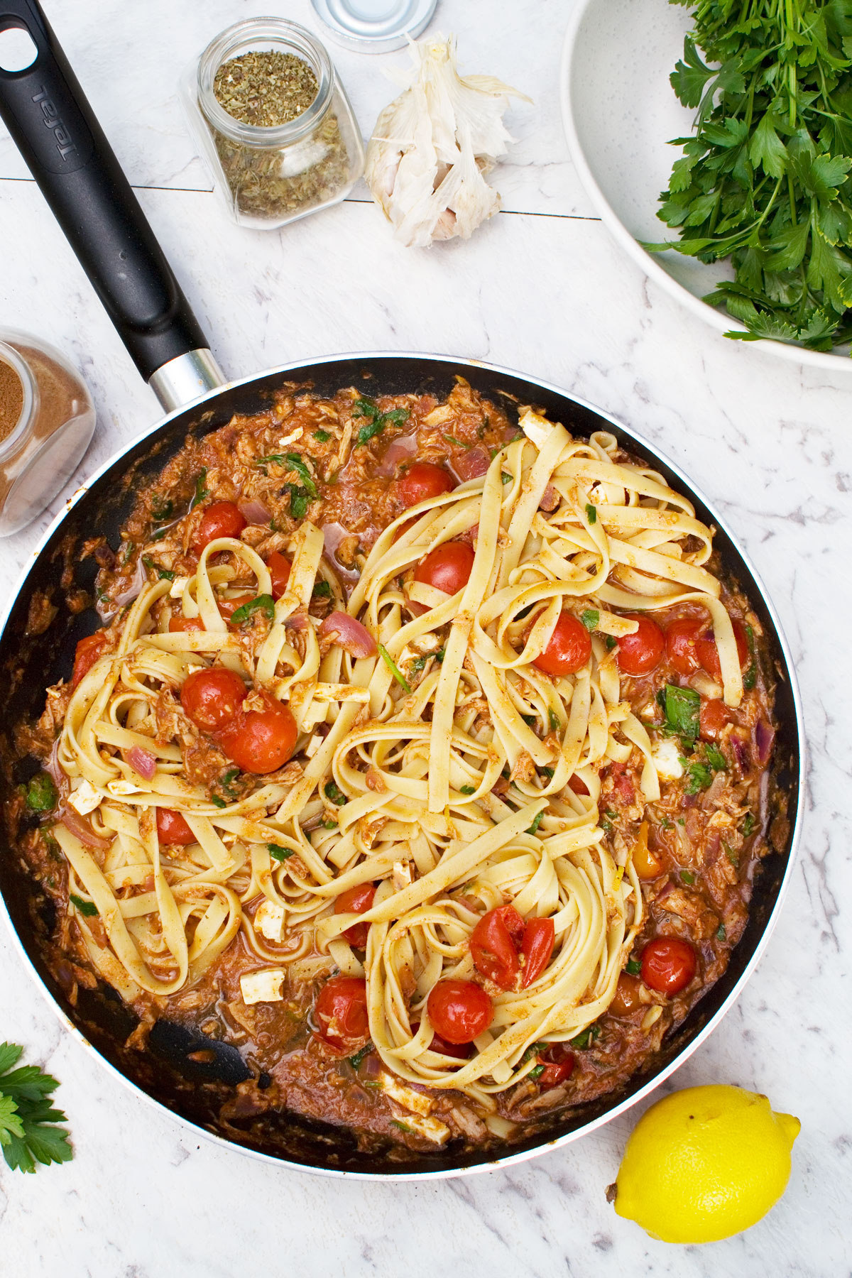 A pan of lemony tuna spaghetti from above on a white marble background. There are a few ingredients scattered around the pan.