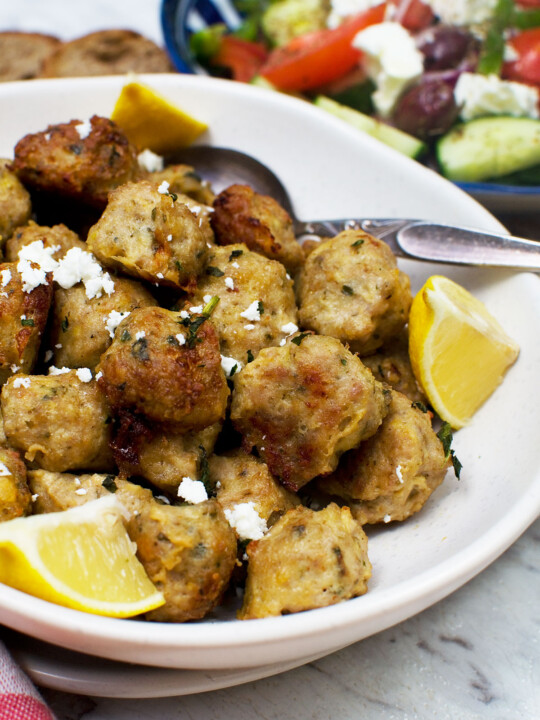 A bowl of Greek air fryer chicken meatballs with slices of lemon and a spoon in - there's also a Greek salad in the background