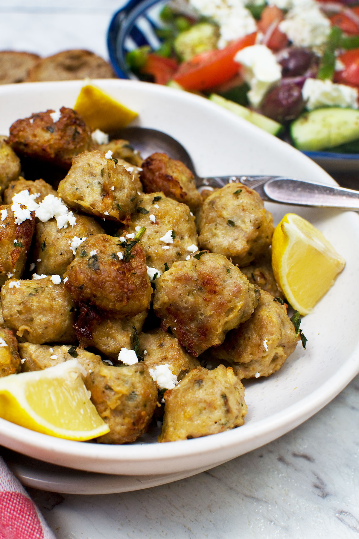 A bowl of Greek air fryer chicken meatballs with slices of lemon and a spoon in - there's also a Greek salad in the background