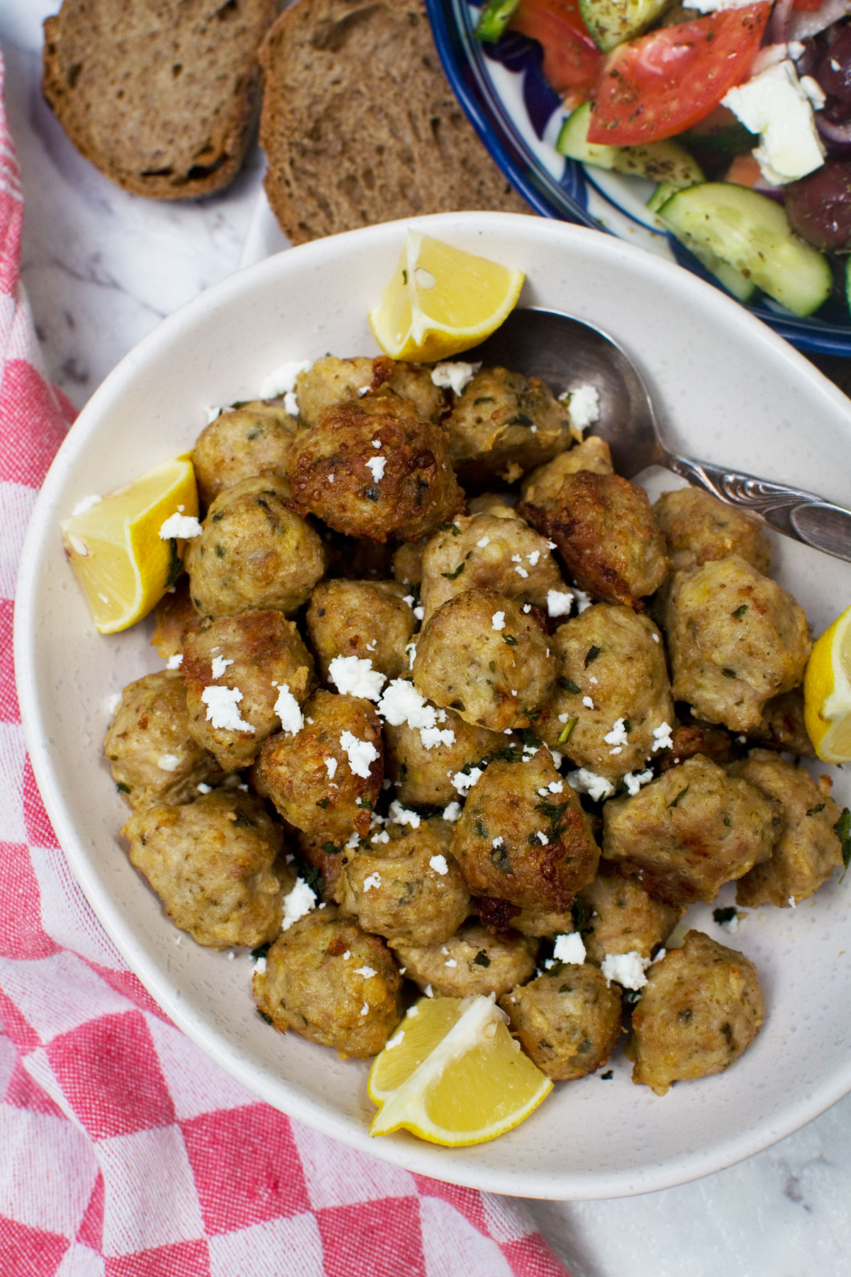 A bowl of Greek air fryer meatballs garnished with lemon wedges and feta and there's a Greek salad at the side