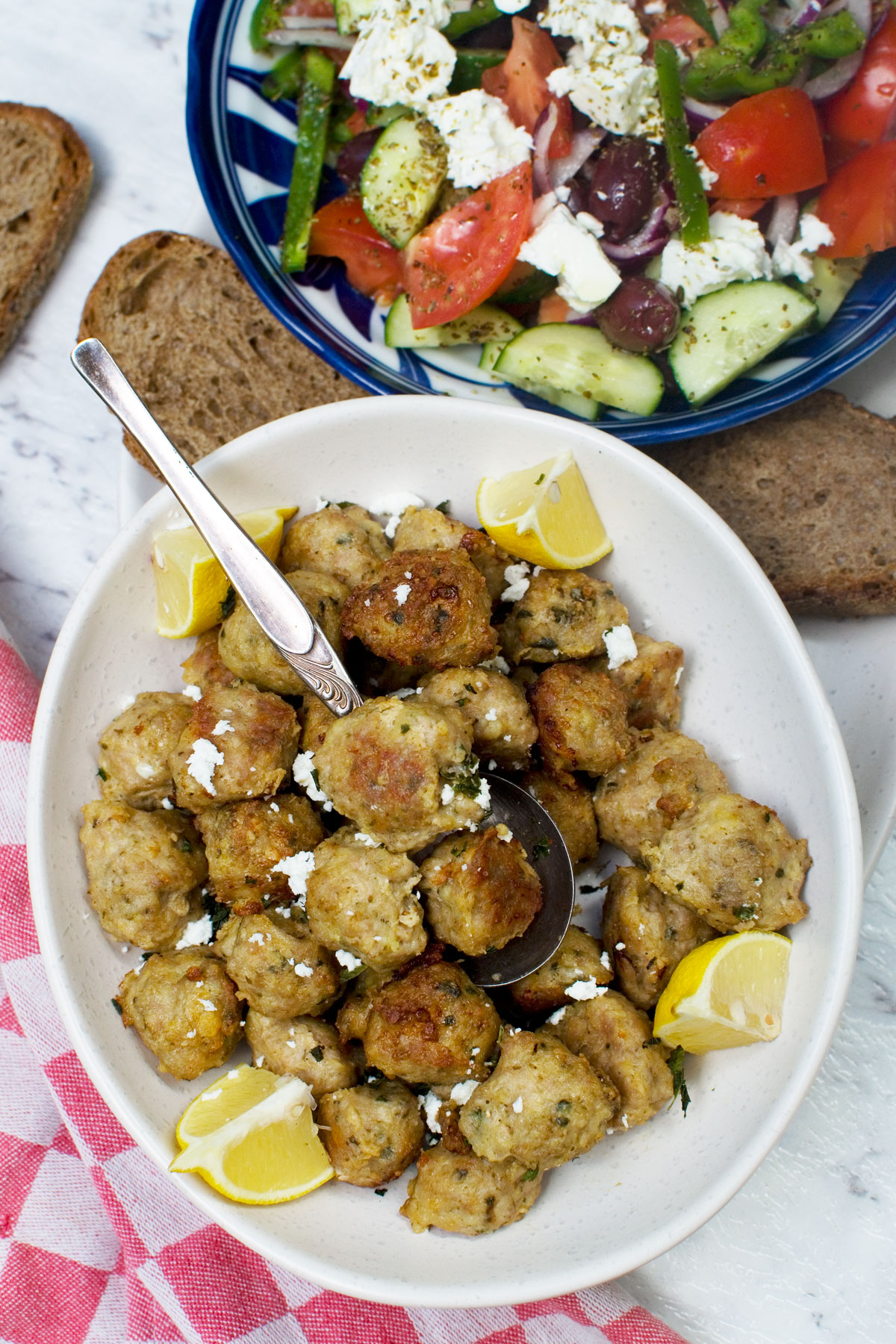 A plate of air fryer Greek meatballs from above with a spoon in and with a Greek salad in the background