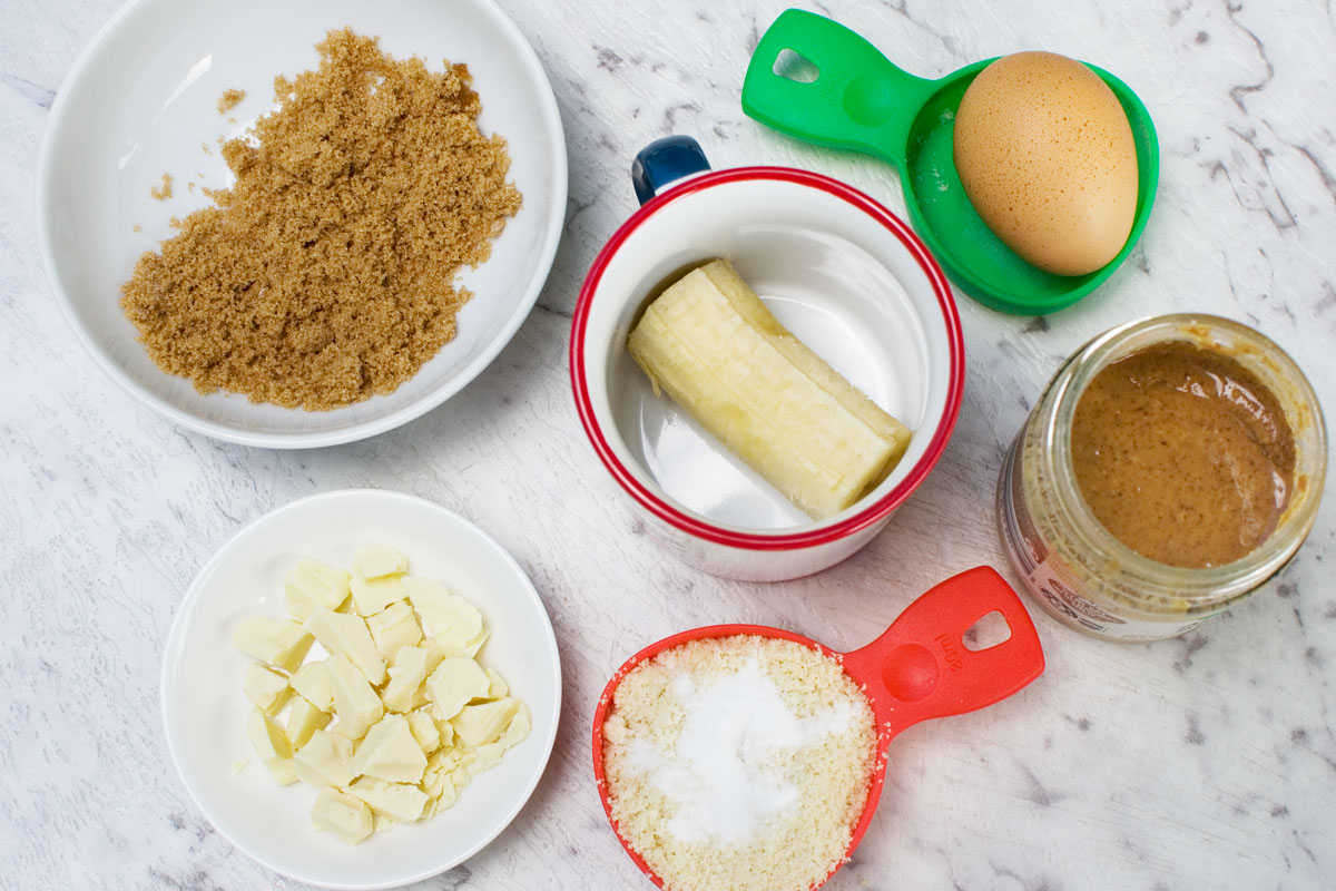 Ingredients to make a gluten free mug cake with banana, almond and white chocolate: brown sugar, 1/2 a banana, white chocolate, almond flour and baking soda, almond butter and an egg