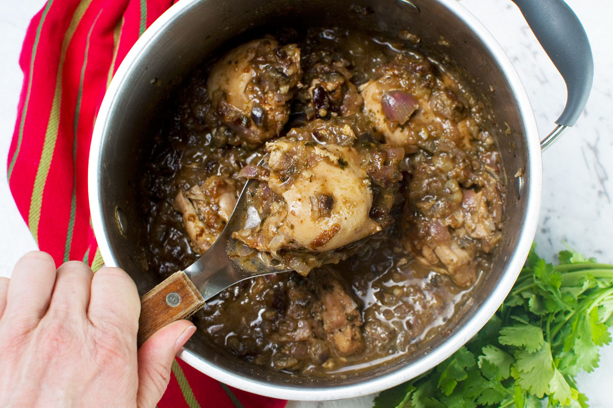 Someone holiding a big spoon of Moroccan chicken stew over the big pot with herbs and a red tea towel in the background