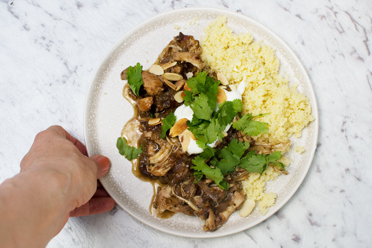 Someone setting a plate of Moroccan chicken stew and couscous down on a marble countertop