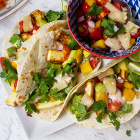 Horizontal image of 3 halloumi tacos with pineapple mango salsa on a white plate with a blue dish of the salsa