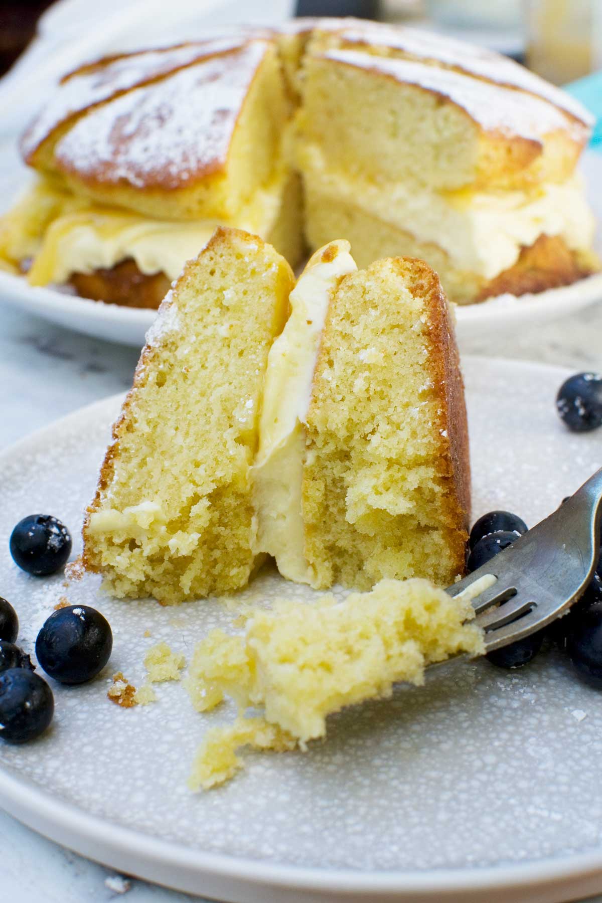 Someone eating a slice of lemon mascarpone cake on a pale beige plate with blueberries with a fork