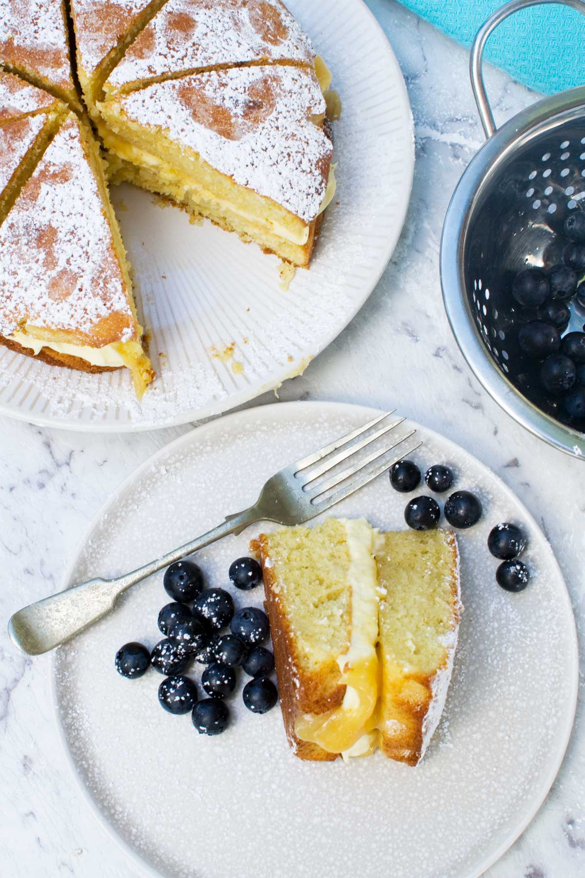 A slice of lemon mascarpone cake on a plate and the rest of the cake on a white plate on a marble background from above