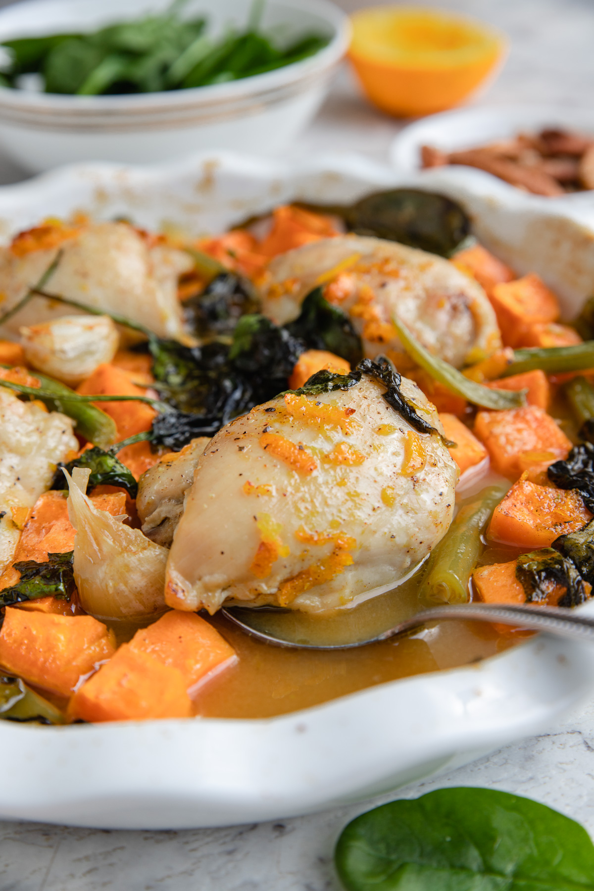 A spoon holding up a chicken thigh in a round baking dish of chicken and sweet potatoes with maple orange sauce