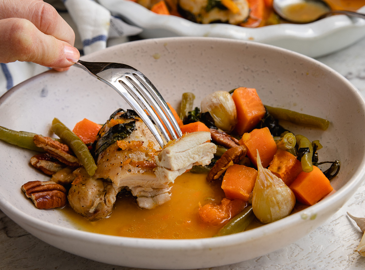 A fork holding up a piece of chicken over a small dish of chicken and sweet potatoes with maple orange sauce