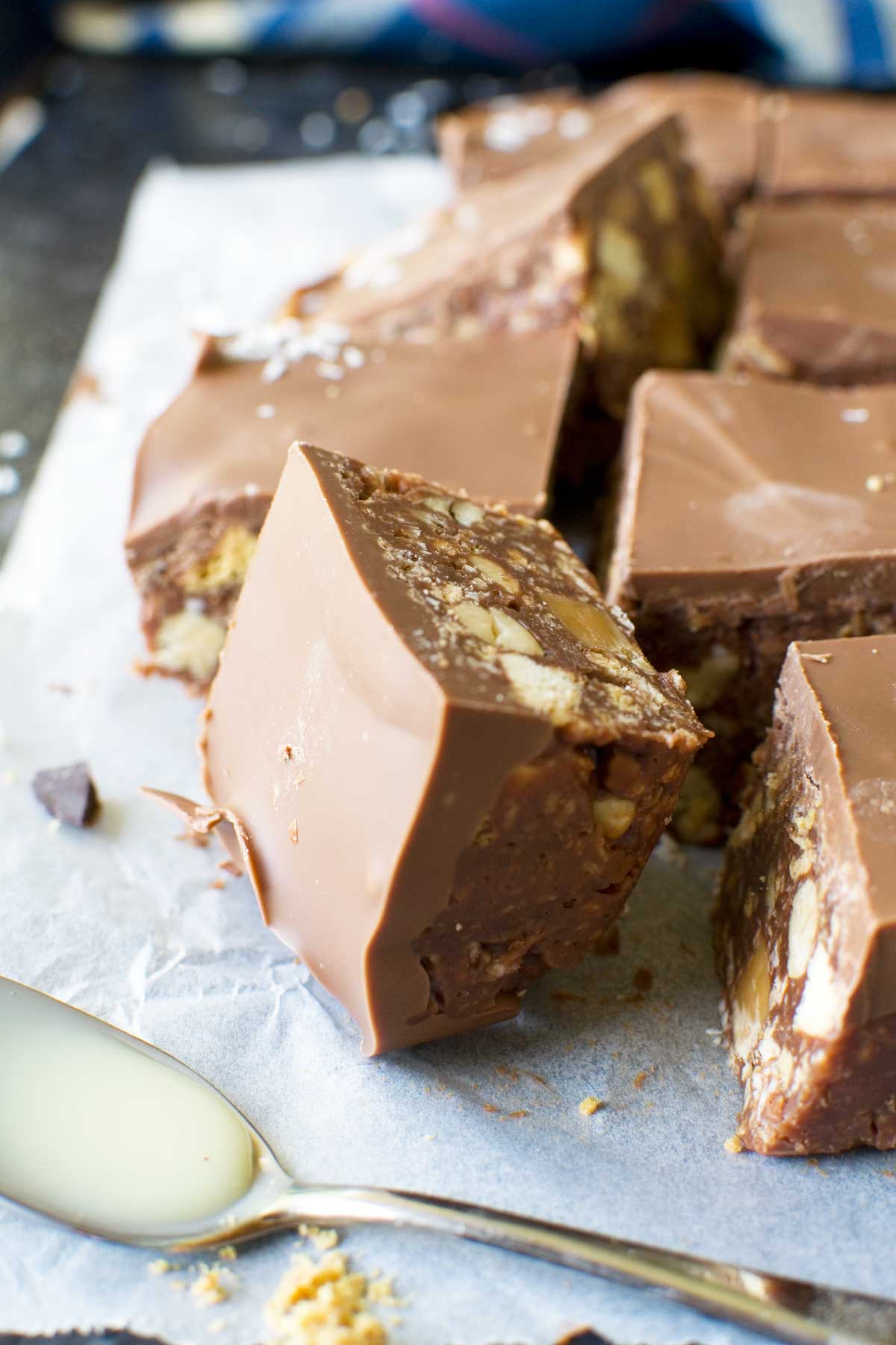 A square of hedgehog slice on its side on baking paper and with more in the background and a spoon with condensed milk in the foreground