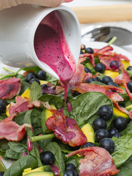 Someone pouring blueberry dressing onto a closeup of a spinach avocado salad