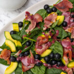 An oval platter of spinach avocado salad with a white jug of blueberry dressing from above on a marble background