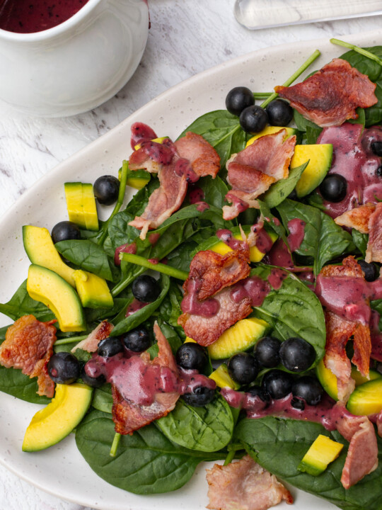 An oval platter of spinach avocado salad with a white jug of blueberry dressing from above on a marble background