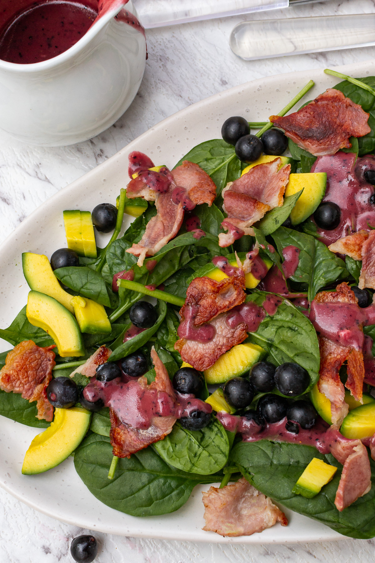 An oval platter of spinach avocado salad with a white jug of blueberry dressing from above on a marble background