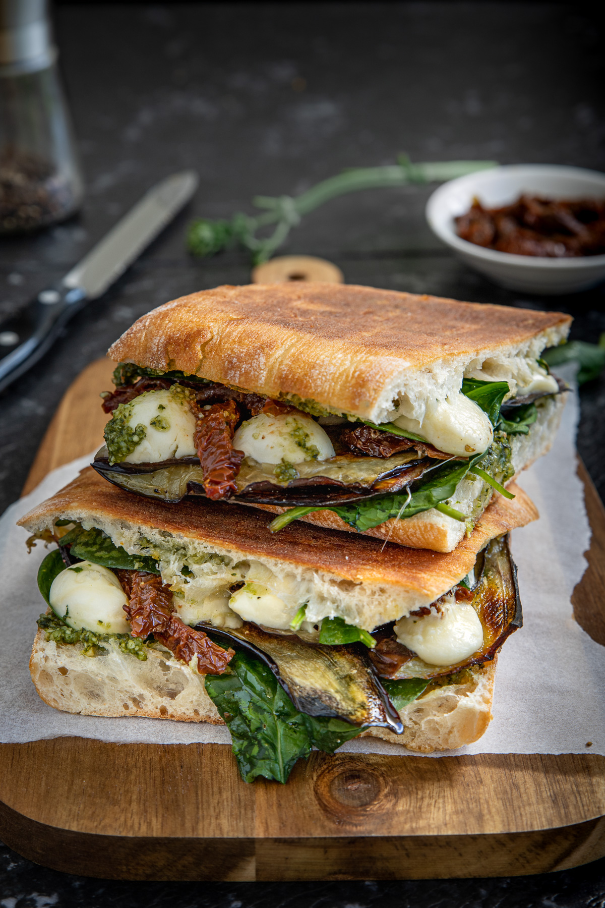 2 toasted veggie sandwiches on top of each other on a wooden board and with ingredients in the background