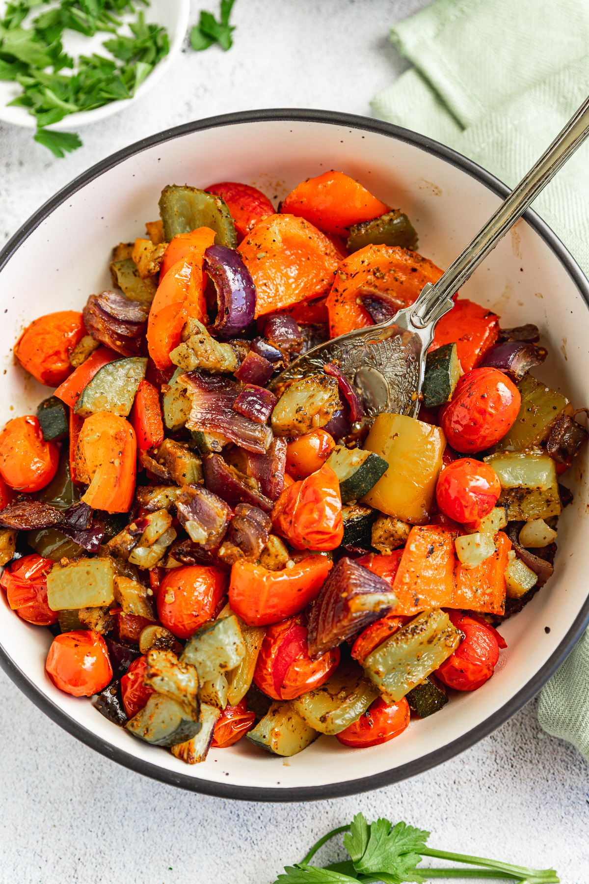 A dish of Mediterranean roast vegetables from above with a spoon in it on a white background and with a green tea towel