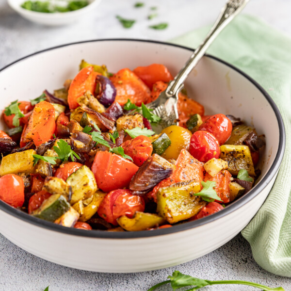 A white dish of Mediterranean roast vegetables on a pale background, with a decorative spoon in it and with a green tea towel and herbs in the background