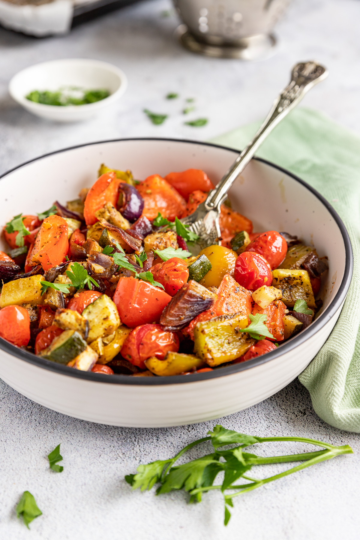 A white dish of Mediterranean roast vegetables on a pale background, with a decorative spoon in it and with a green tea towel and herbs in the background