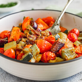 A dish of Mediterranean roast vegetables on a white background with a spoon in it and a green tea towel in the background