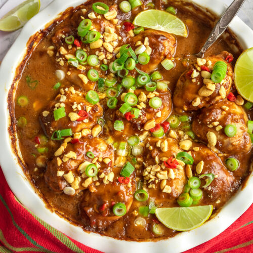 A white baking dish of spicy peanut butter chicken from above with a red and green striped tea towel and ingredients around it