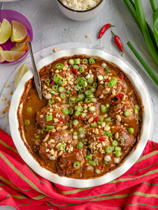 A round white baking dish of spicy peanut butter chicken from above with a stripy red and green tea towel and ingredients around the dish