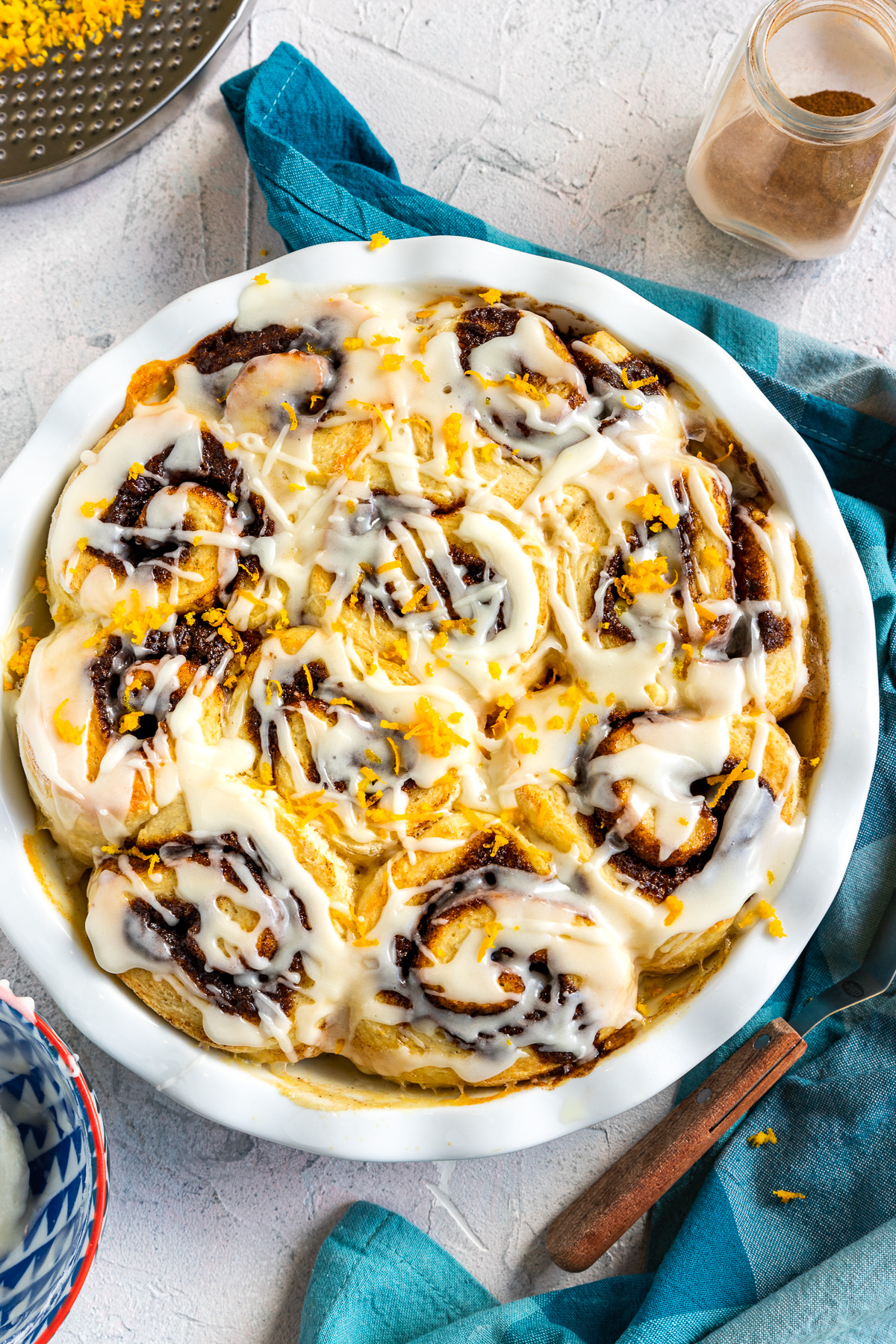 A round white baking dish of cinnamon scrolls from above on a white background and with a blue checked tea towel around it