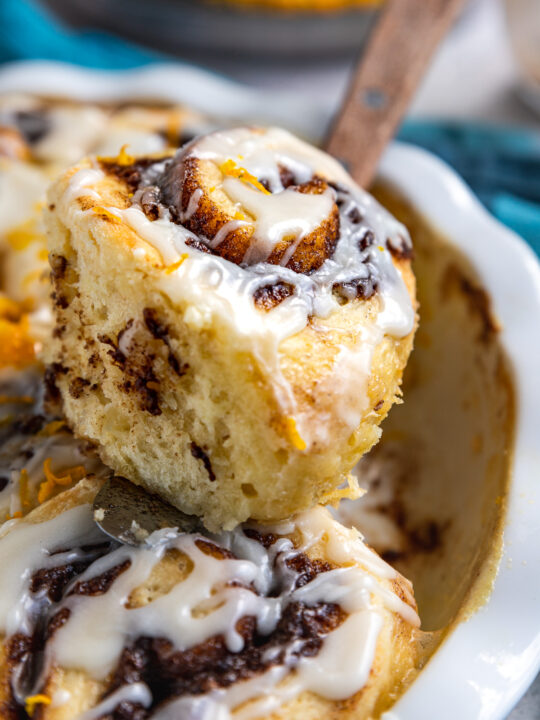 Closeup of someone taking a cinnamon scroll out of the pan with a wooden handled palette knife