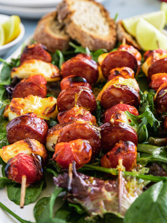 A closeup of 4 halloumi kebabs on a white platter and on a bed of green leaves with bread and lime segments on the side