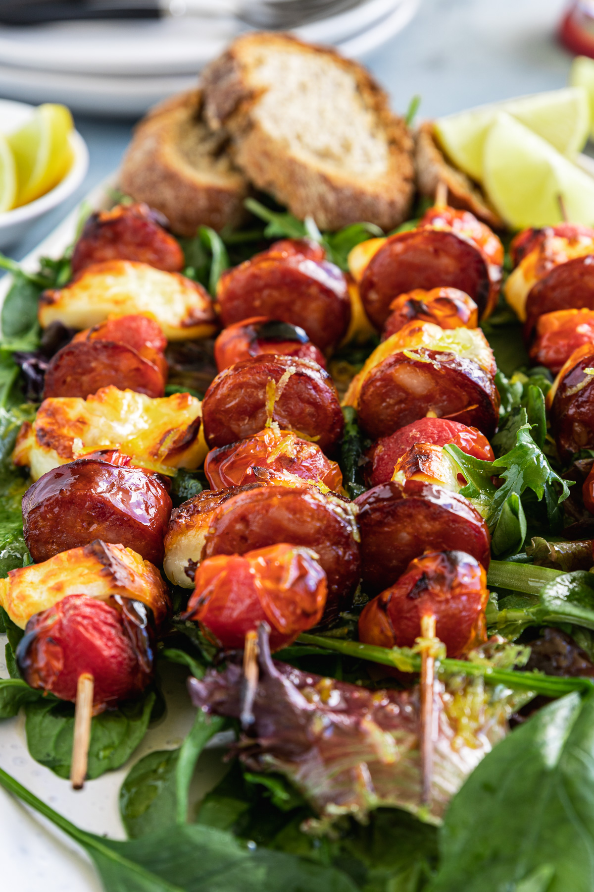 A closeup of 4 halloumi kebabs on a white platter and on a bed of green leaves with bread and lime segments on the side