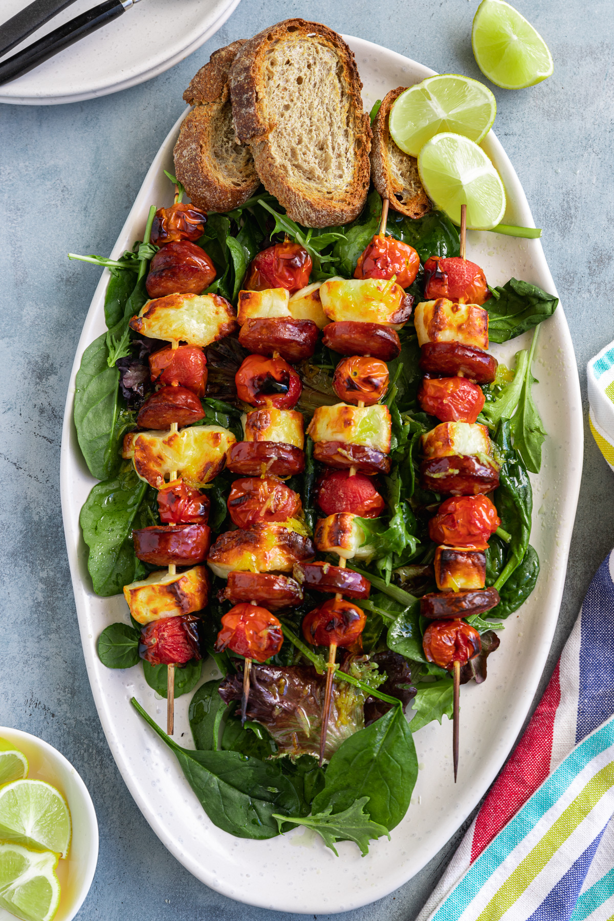 A platter of 4 halloumi kebabs on a white platter on a bed of salad leaves with grilled bread and lime wedges and with a multi-coloured tea towel at the side