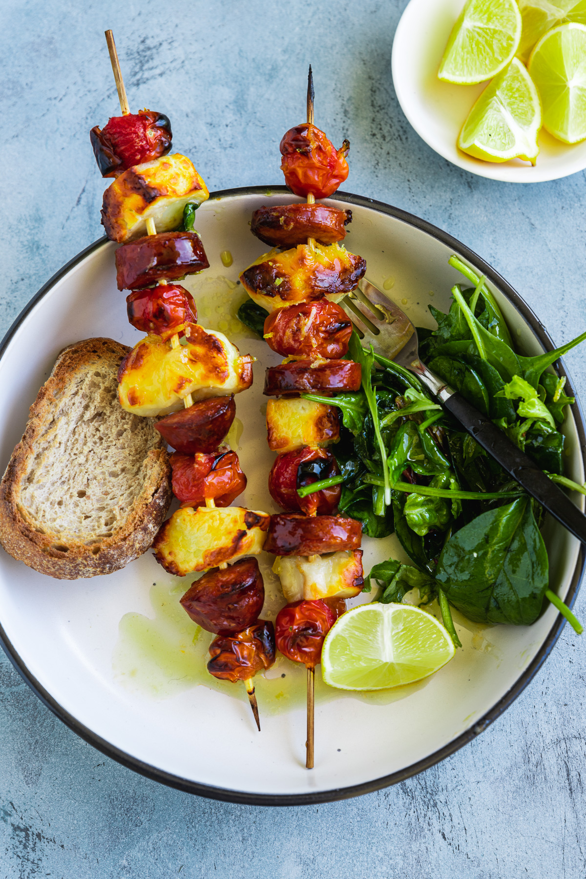 A dish of 2 halloumi kebabs with chorizo and tomatoes from above served with toasted bread, green salad leaves and lime wedges on the side