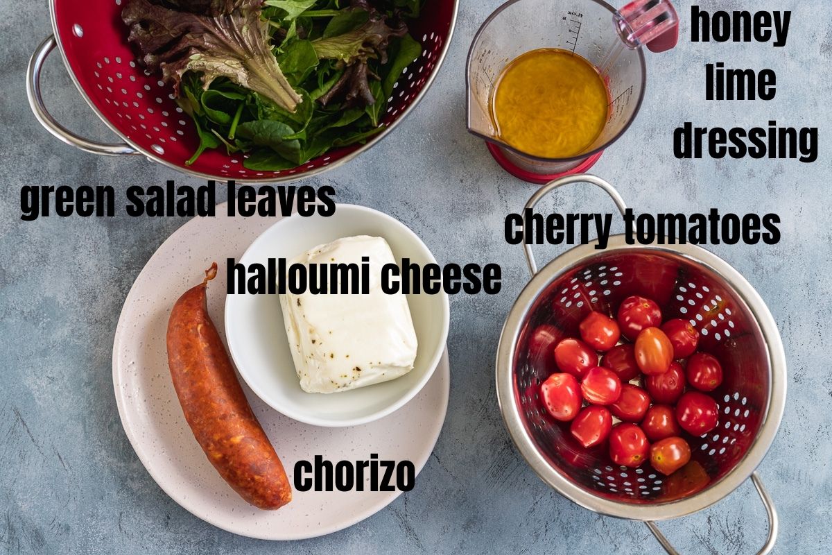 All the ingredients you need to make halloumi kebabs with chorizo and tomatoes including green salad leaves, honey lime dressing, halloumi cheese log, chorizo sausage and cherry tomatoes