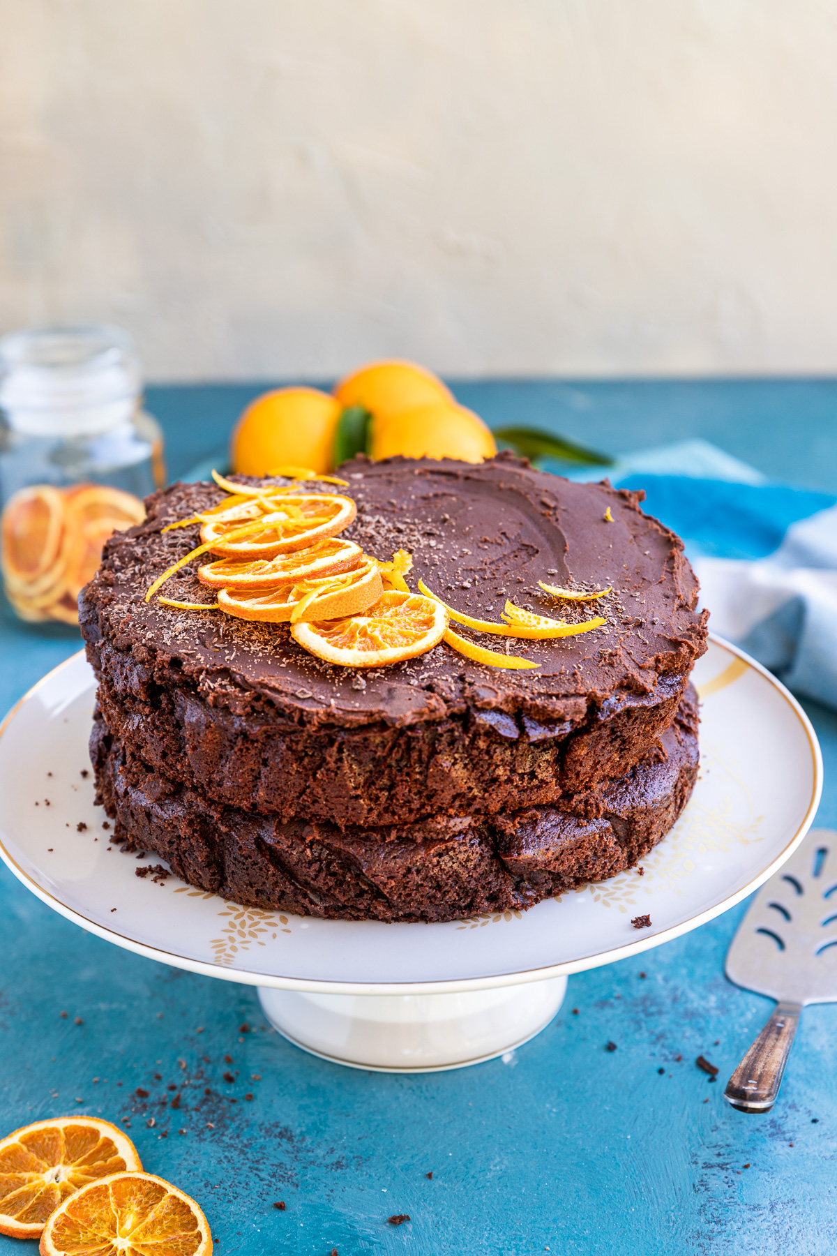 A finished chocolate orange cake decorated with orange slices on a white cake stand and on a blue background with a grey wall and ingredients behind