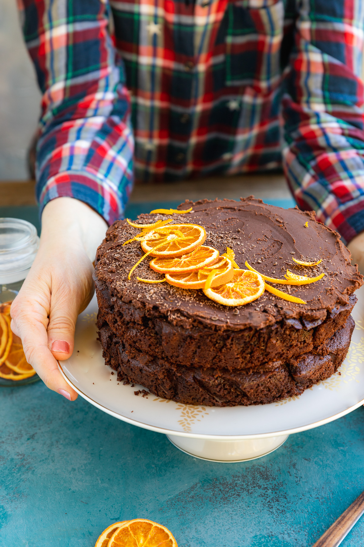 Someone in a red and blue checked shirt placing a decorated chocolate orange layer cake on a white cake stand on a blue surface