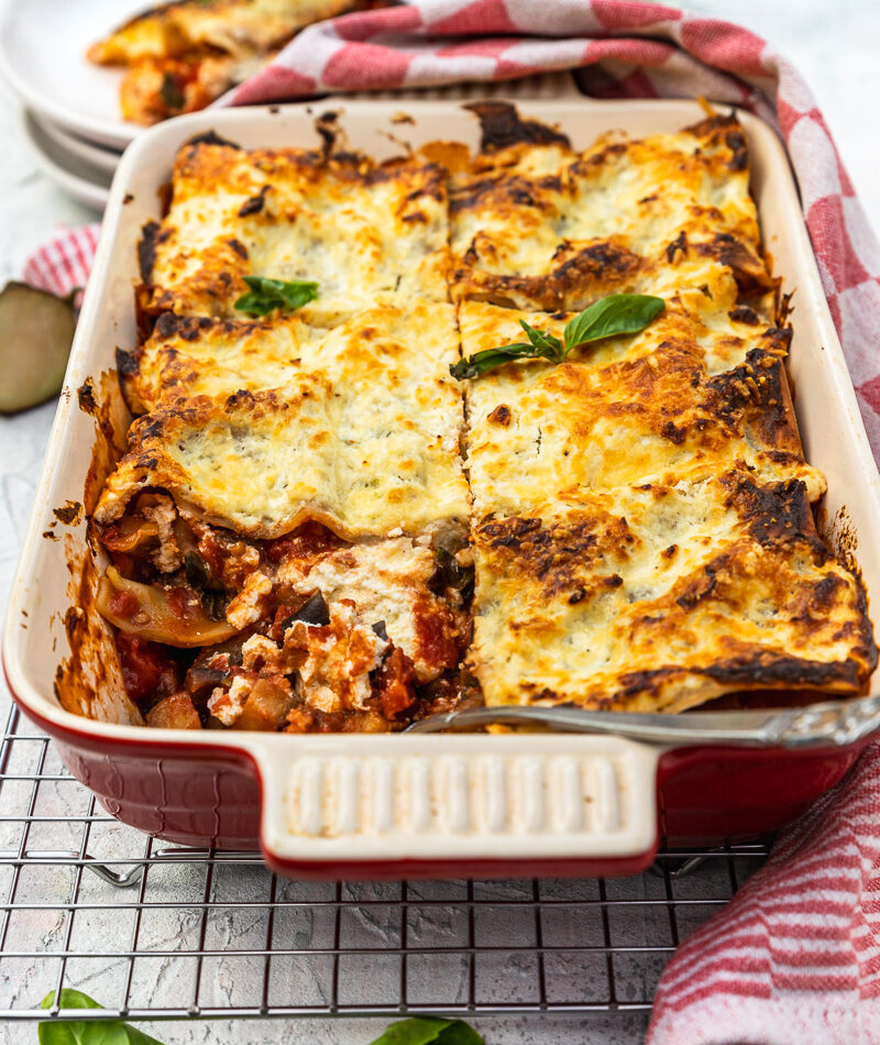 A red baking dish of eggplant lasagna with a piece missing on a cooking rack and with a red checked tea towel in the foreground