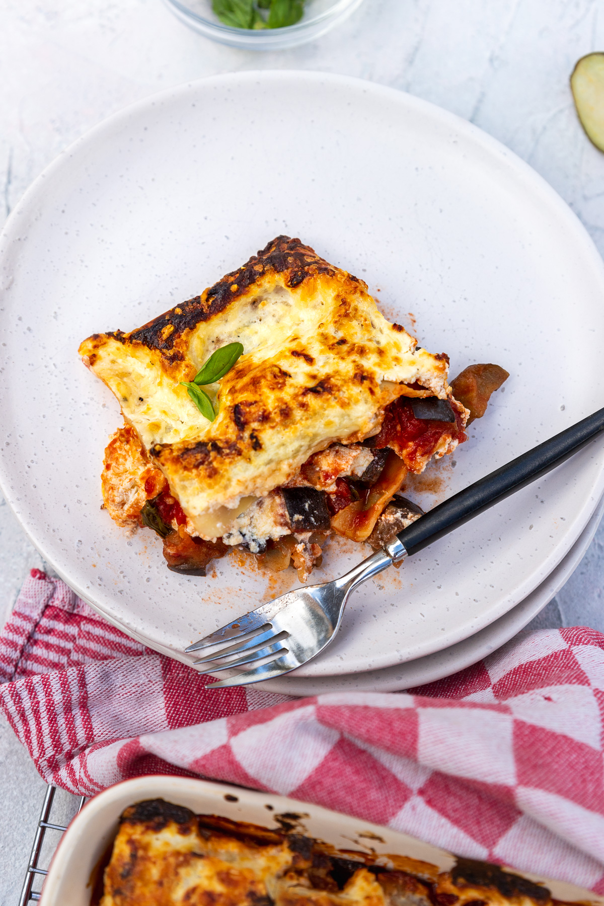 A piece of eggplant lasagna from above on a white plate with a red checked tea towel and the edge of the baking dish