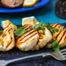 A small black slate platter of cook halloumi with basil and lemon wedges and a fork and plate of toasted bread in the background