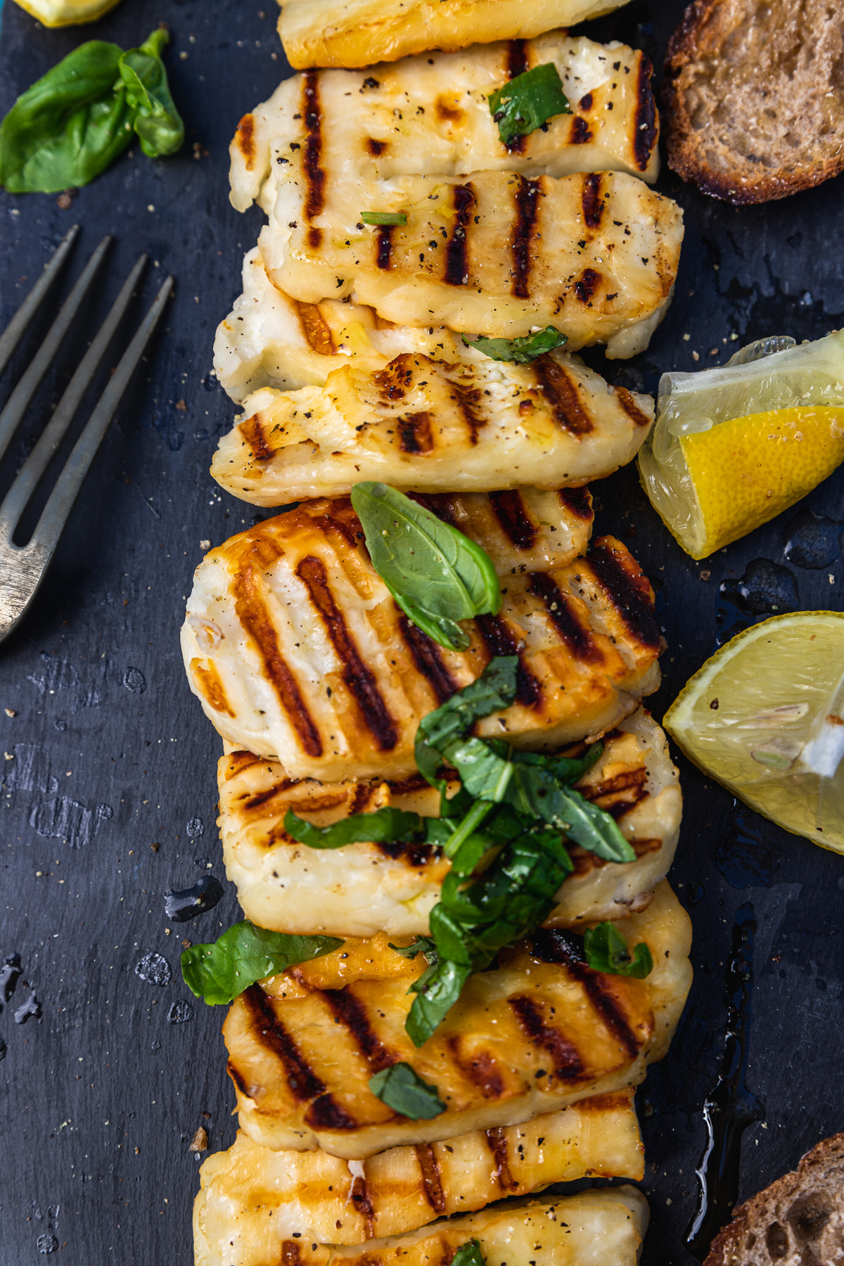 Slices of grilled halloumi on a black slate platter from above with lemon segments, basil and a fork