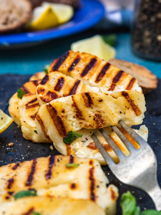 Closeup of someone grabbing a piece of grilled halloumi with a fork from a black platter with more in the background