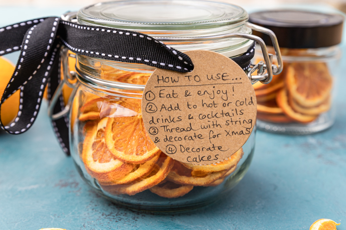 A jar of dried oranges on a blue background and with a black ribbon around the top and a brown label with ways to use the oranges on