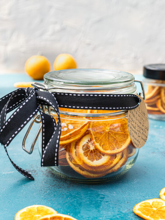 A mason jar full of dried orange slices with a black bow around it and brown label on a blue table top and with oranges and a white wall in the background