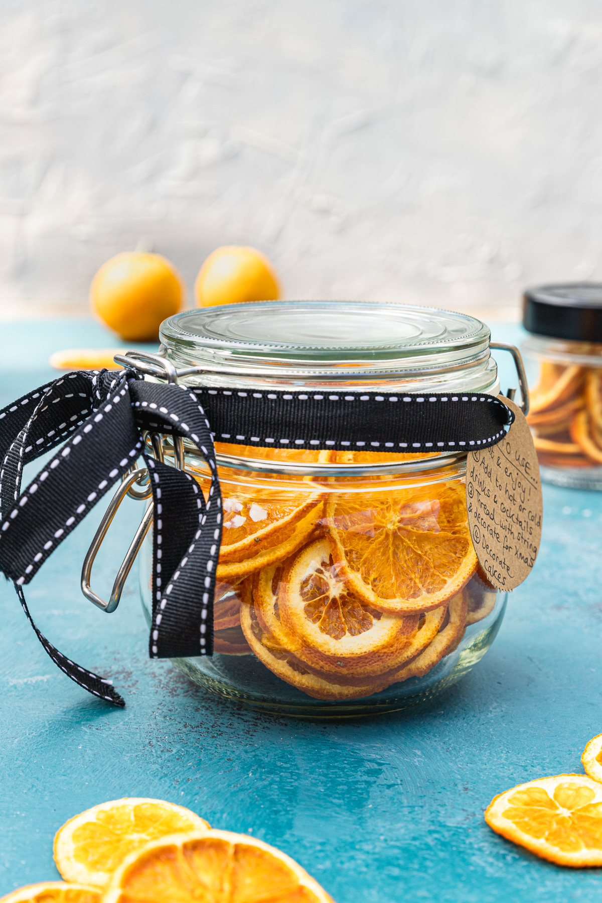 A mason jar full of dried orange slices with a black bow around it and brown label on a blue table top and with oranges and a white wall in the background