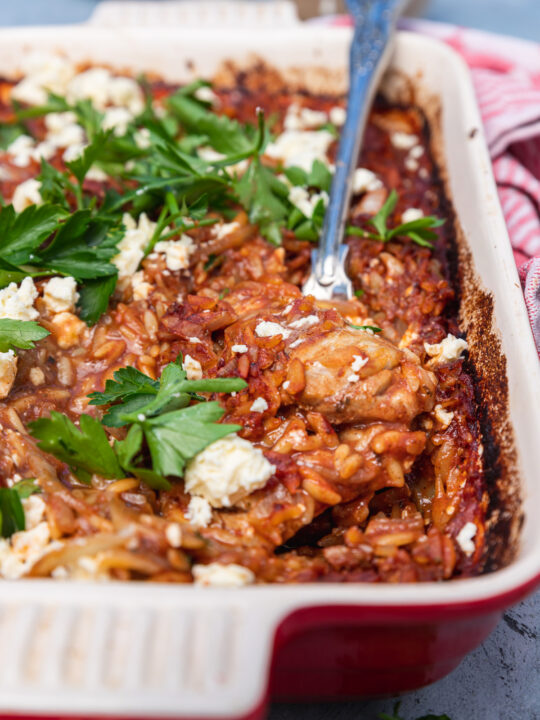 A red baking dish of Greek giouvetsi with chicken with parsley and feta on it and a spoon in it and with a red checked tea towel in the background