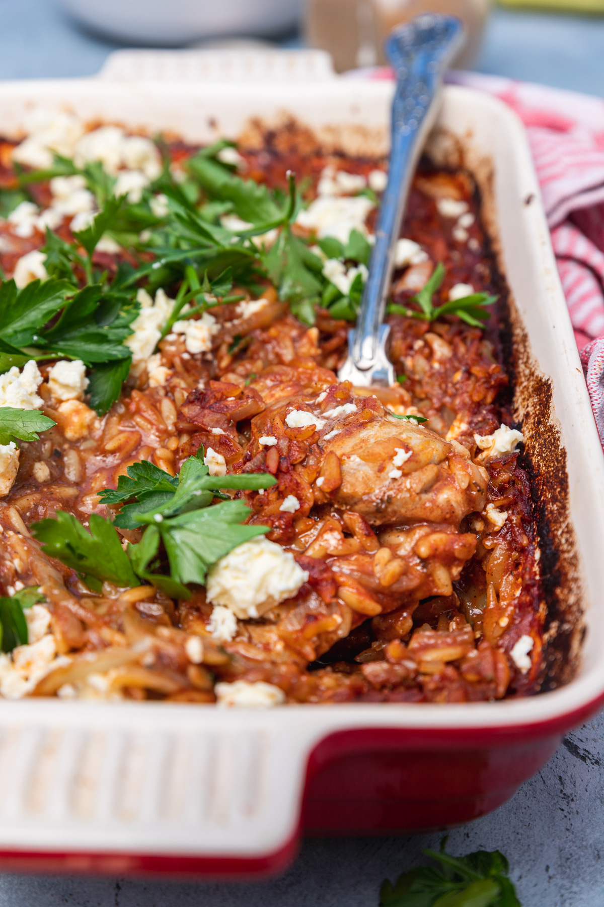 A red baking dish of Greek giouvetsi with chicken with parsley and feta on it and a spoon in it and with a red checked tea towel in the background