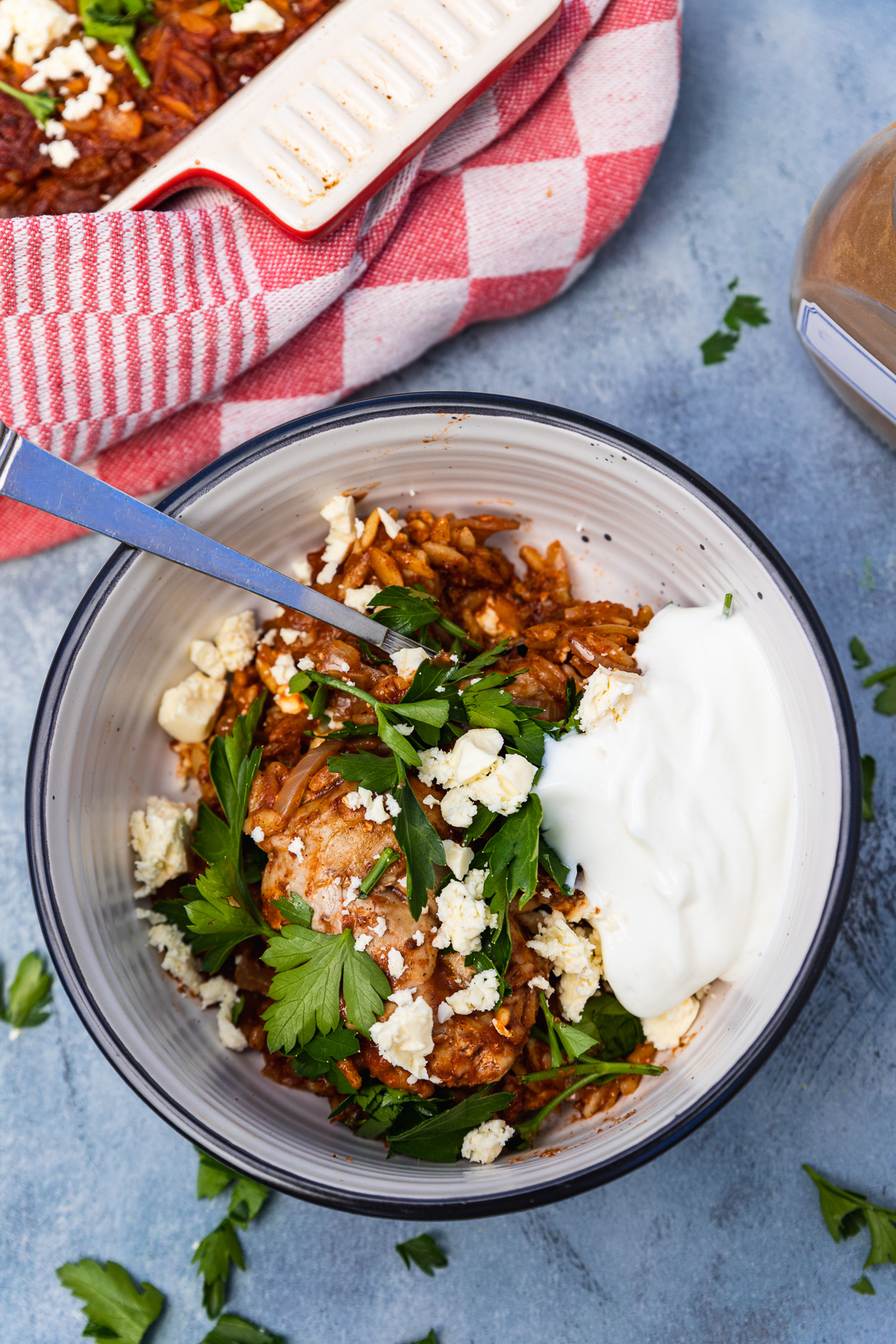 A bowl of giouvetsi with chicken with a spoon in it and with some Greek yogurt from above and on a blue background with a baking dish in the background