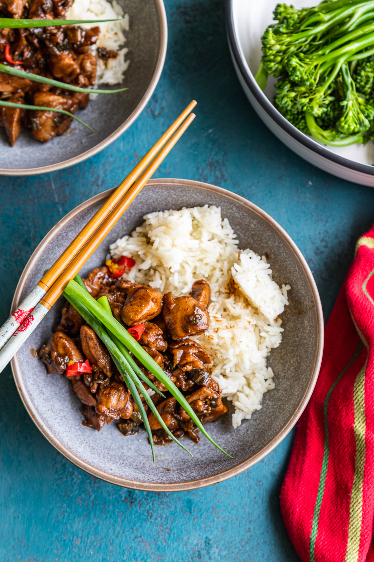 A dish of Shanghai chicken with chopsticks laid over the top from above on a blue background and with a red stripy tea towel