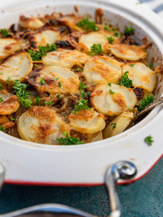 A closeup of a baking dish of boulangere potatoes or potato bake with bacon with a blue and white stripe tea towel in the background