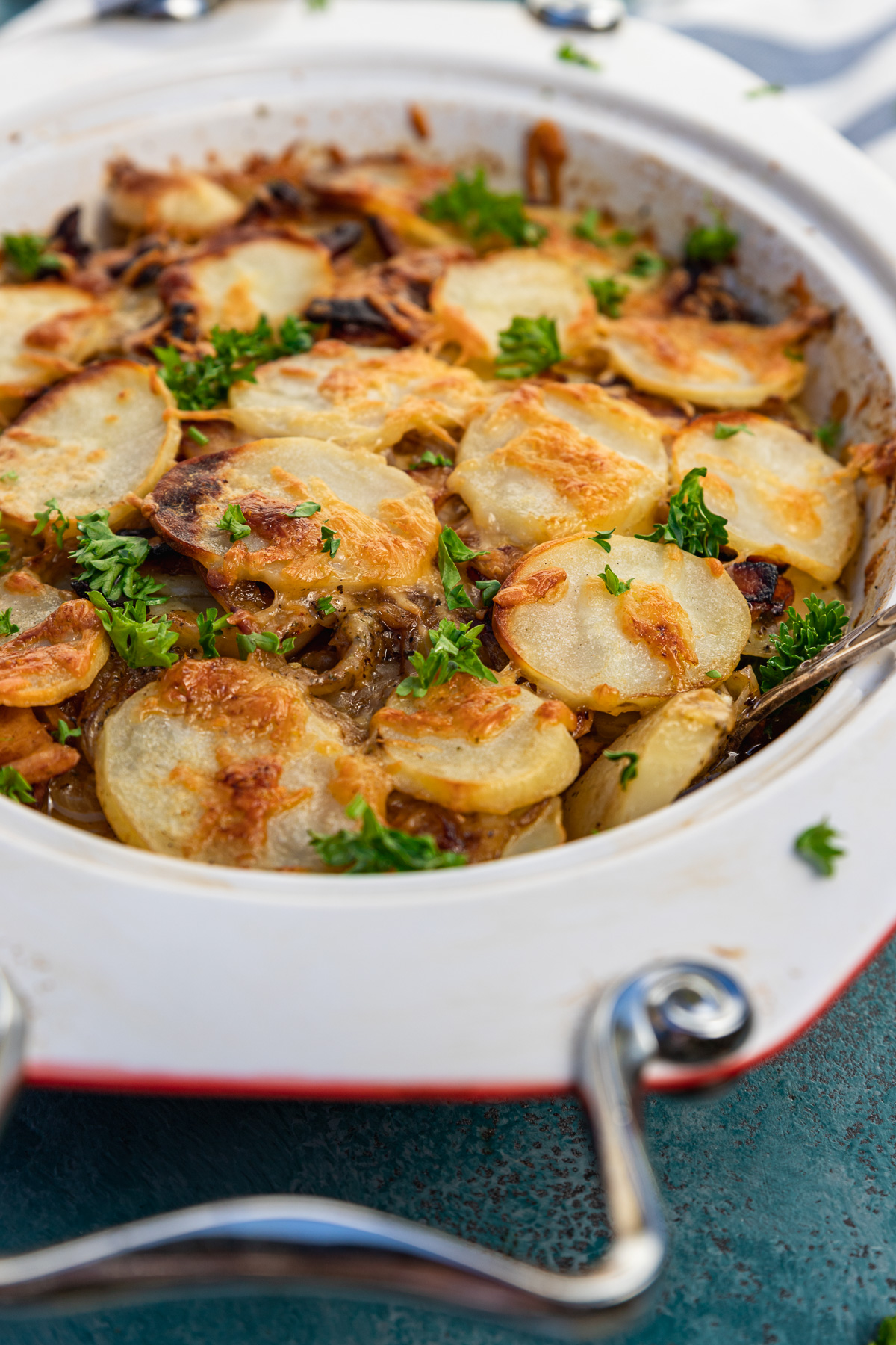 A closeup of a baking dish of boulangere potatoes or potato bake with bacon with a blue and white stripe tea towel in the background