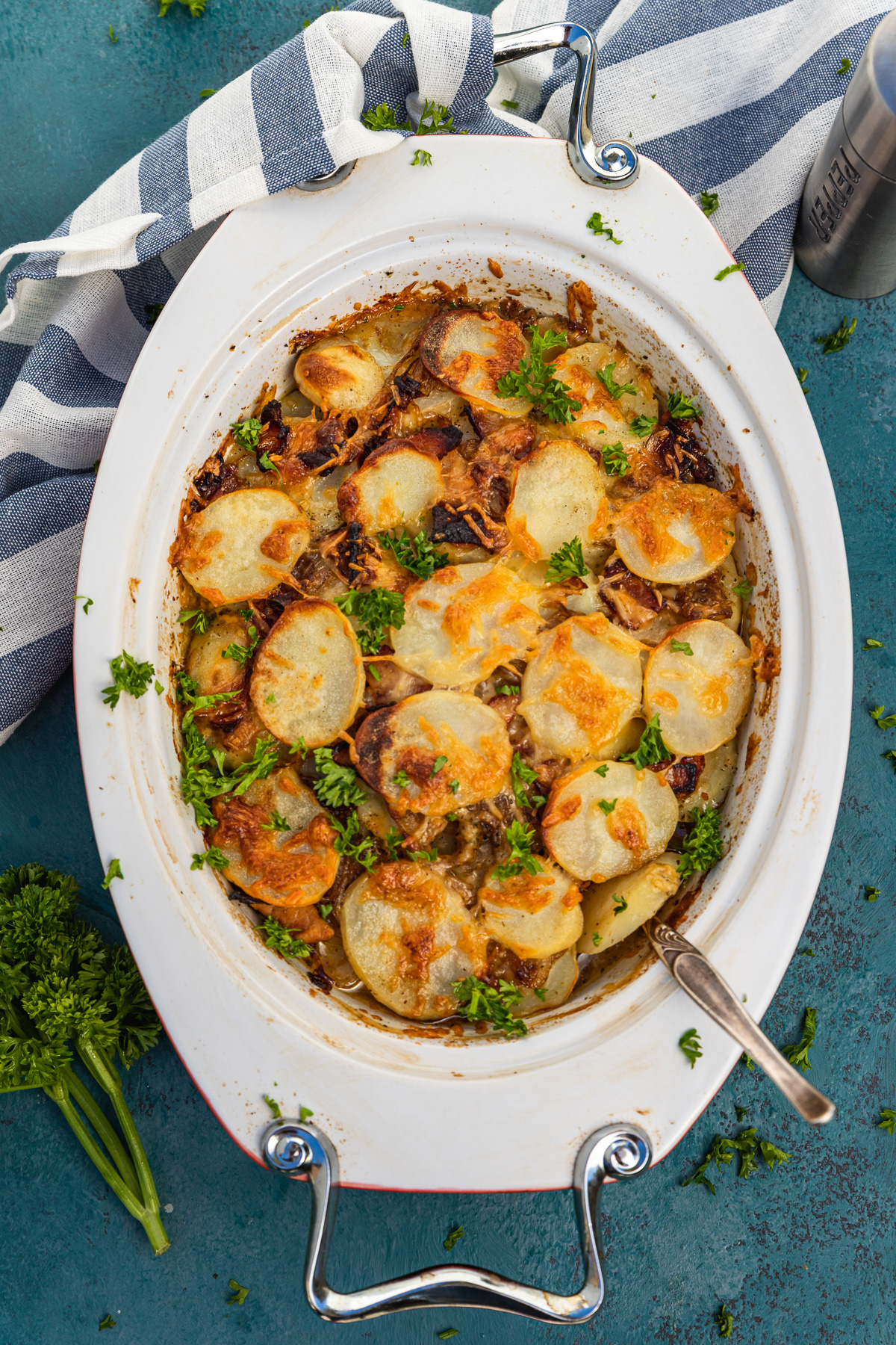 An oval-shaped baking dish of potato bake with bacon from above on a dark blue background and with a blue and white striped tea towel